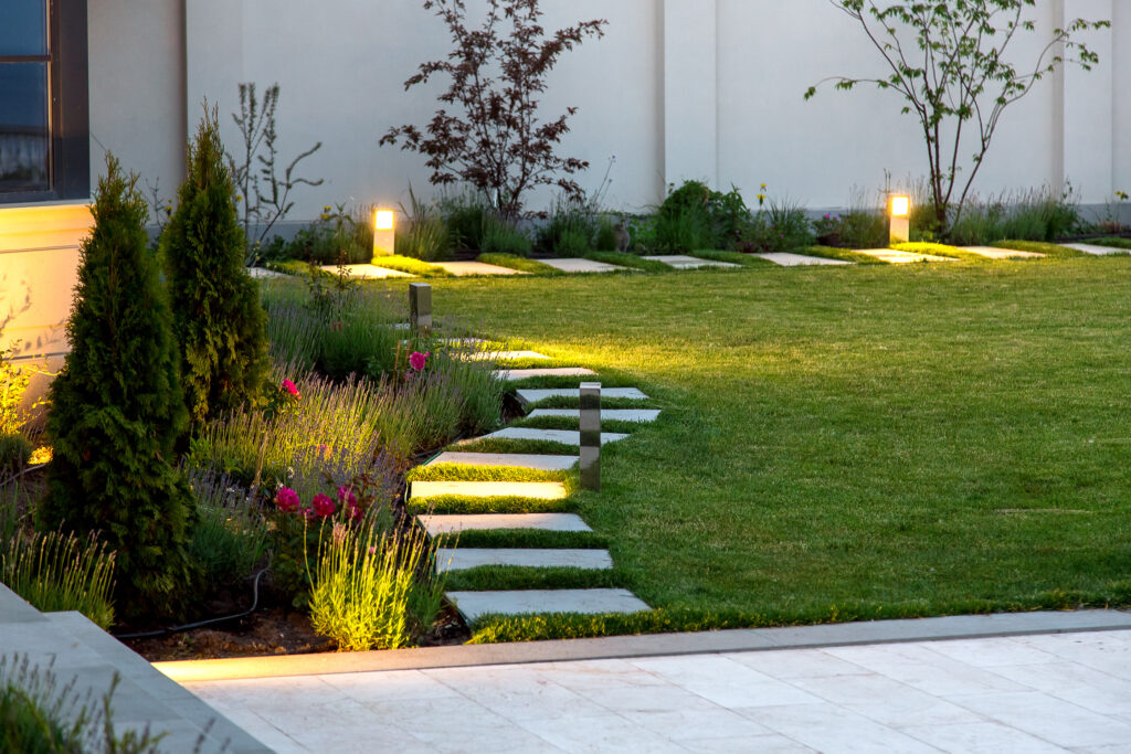 A well-lit outdoor garden pathway featuring modern step lights, surrounded by lush greenery and flowers in a neatly landscaped yard.