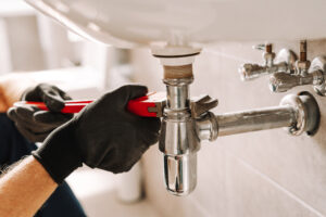 Binsky Home Plumber in black gloves fixes a sink pipe with a wrench in a bathroom, showcasing precision and craftsmanship